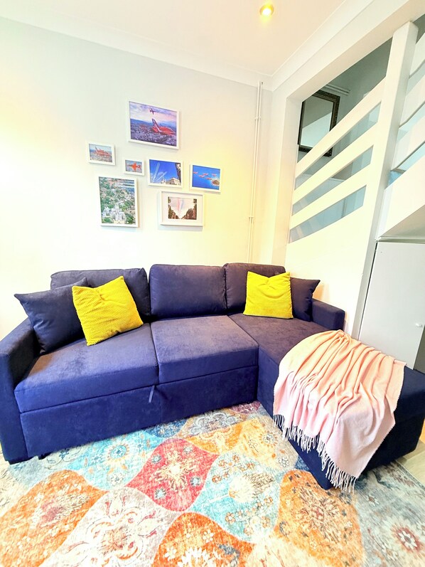 Una sala de estar con un sofá azul, cojines amarillos, un tapete colorido y una escalera con barandillas blancas.