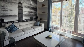 A modern living room with a grey sofa, a white coffee table, a mural of a lighthouse, and a large window with curtains. - Bora Premium by Grand Apartments (Sopot)