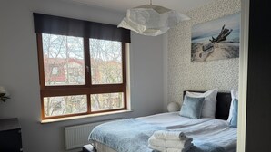 A bedroom with a large window, a bed with a blue quilt, a white radiator, and a wall-mounted painting. - Bora Premium by Grand Apartments (Sopot)