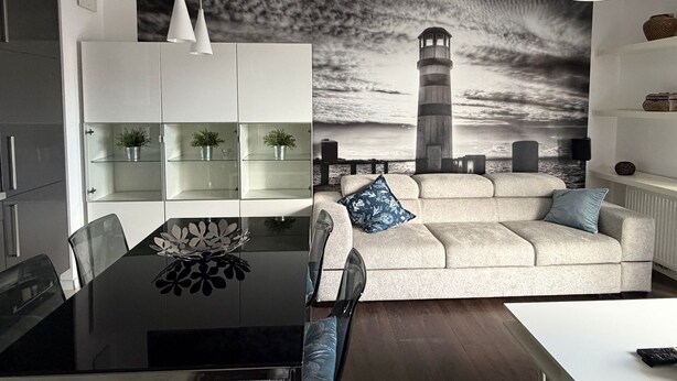 A modern living room with a large wall mural, a dining table with chairs, a sofa with cushions, and a white ceiling light. - Bora Premium by Grand Apartments (Sopot)