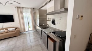A modern kitchen with a built-in oven, a sink, and a stove. There is a beige sofa with decorative pillows and a flat-screen TV mounted on the wall.