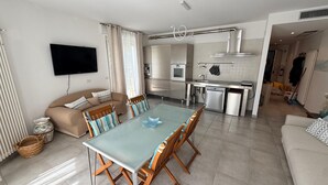 Een moderne woonkamer met een bank, een eettafel met stoelen, een keuken met apparatuur en een wandgemonteerde flatscreen-tv.