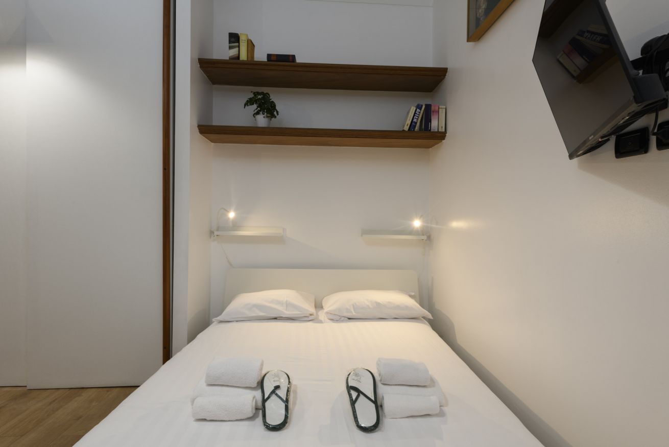 A modern hotel room with a bed, white linens, a small plant on a shelf, and a TV mounted on the wall.