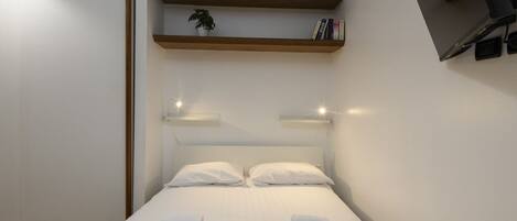 Habitación de hotel moderna con cama, ropa de cama blanca, una planta pequeña en un estante y un televisor montado en la pared.