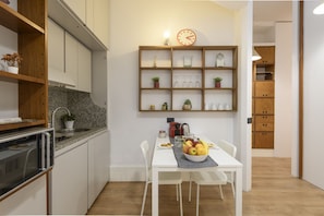 Une cuisine moderne avec des armoires blanches, un comptoir en granit, un four encastré, une table à manger avec des chaises et une étagère murale où sont exposés divers objets.