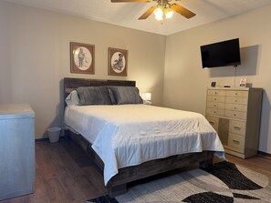 A bedroom with a wooden bed, a nightstand with a lamp, a dresser, a television, and two framed pictures on the wall.