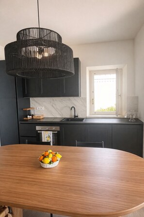 Et moderne køkken med mørke skabe, en marmorbeklædt bagvæg, et træspisebord med en skål frugt, og en sort hængende lampe.