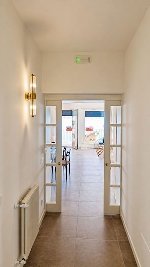 Un couloir avec un radiateur, un luminaire et une porte donnant sur un balcon aménagé avec du mobilier d’extérieur.