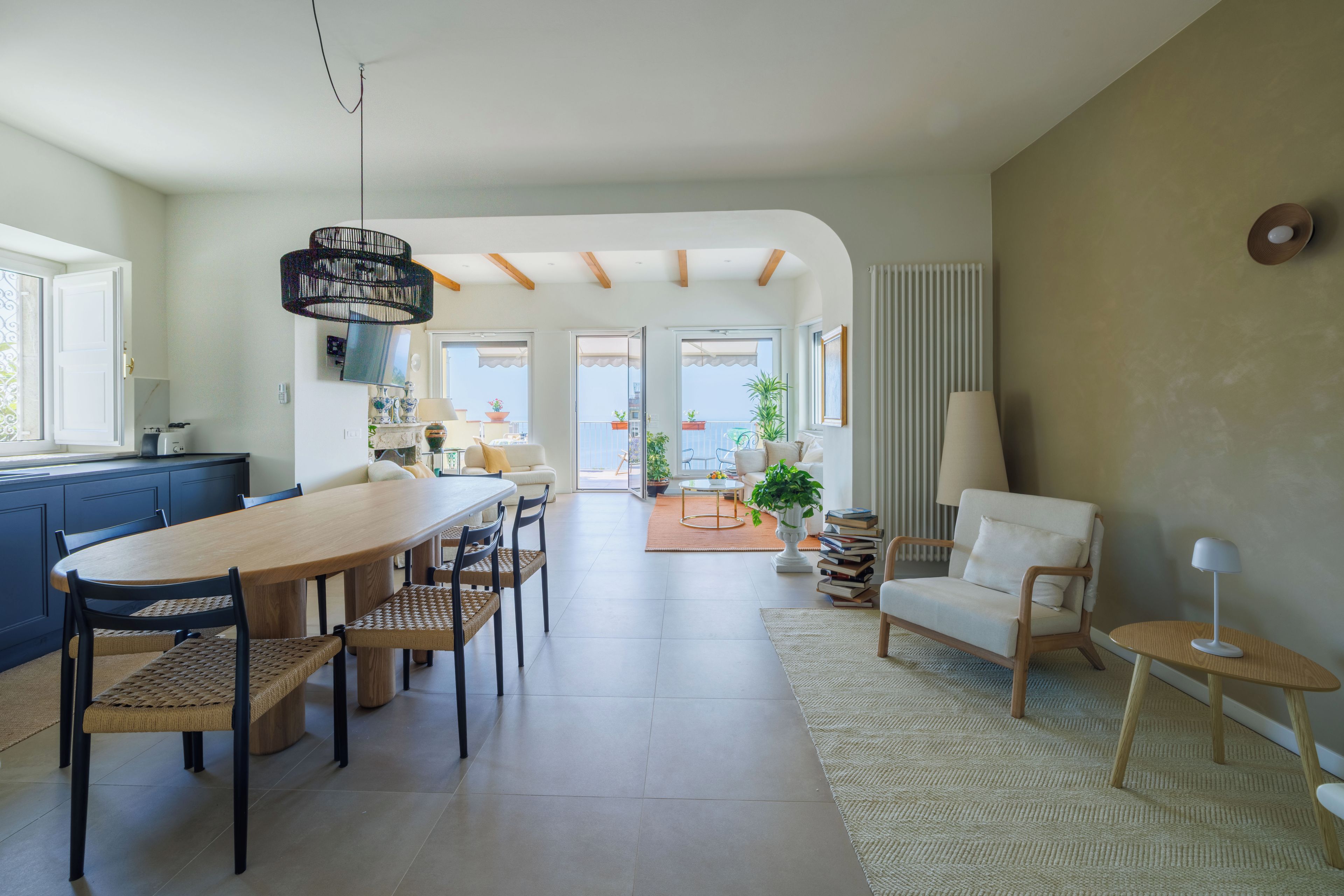 A spacious living area with a dining table, chairs, and a sofa. There is a kitchen area with cabinets and a countertop. The room has large windows and a view of the outdoors.