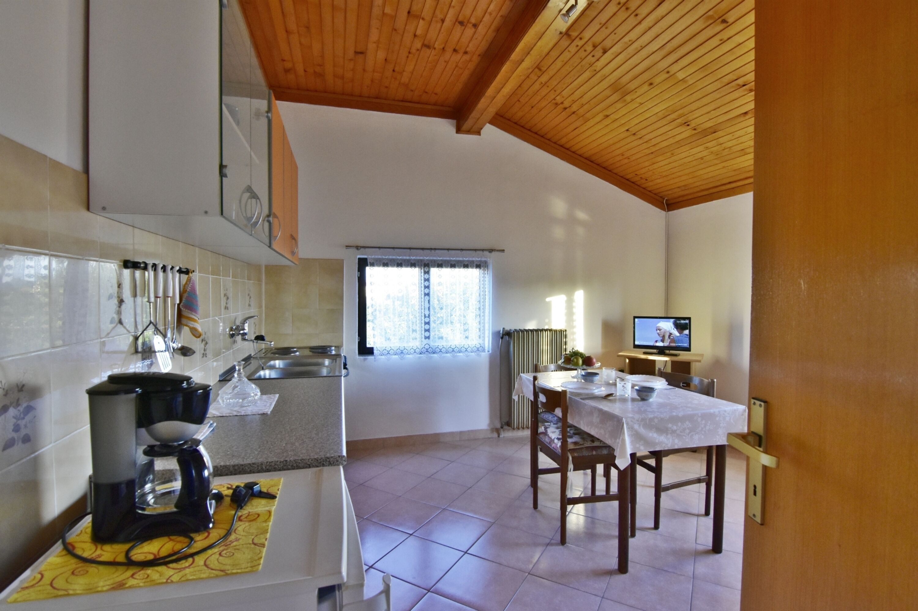 A kitchen with a coffee maker, a dining table with chairs, a television, and a window with curtains.