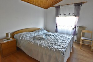 Un dormitori amb un llit, una nit, una cadira, una finestra amb cortines i un sostre de fusta.