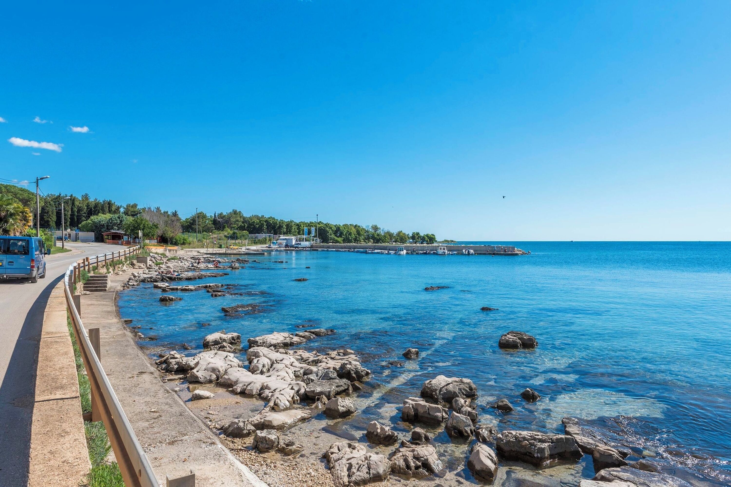 Una carretera costera con un litoral rocoso, un cielo azul despejado y una vista distante de un muelle.