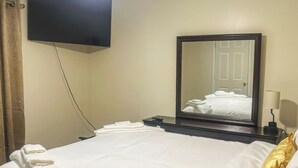 Ett hotellrum med en säng, en platt-TV som är monterad på väggen, ett nattduksbord med en lampa och en stor spegel som reflekterar rummet.