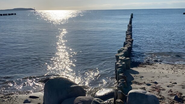 A coastal scene with a long, narrow breakwater extending into the water, a sandy beach with scattered rocks, and a distant landmass. - Holiday Home in Dranske (Dranske)