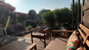 A wooden deck with a sofa and chairs, a metal railing, and a view of greenery and a building in the background. - Villa Roccapina (Sotta)