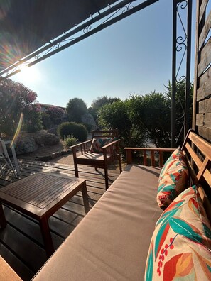 Eine Holzterrasse mit einer Couch und Sesseln, einem Metallgeländer und Blick auf Grünflächen sowie ein Gebäude im Hintergrund.