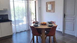 A modern living room with a sofa, a dining table with chairs, a small round table with a candle, and a kitchen area with a refrigerator and a window. - Villa Roccapina (Sotta)