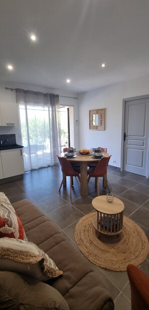 Ein modernes Wohnzimmer mit einer Couch, einem Esstisch mit Stühlen, einem kleinen runden Tisch mit Kerze und einer Küchenzeile mit Kühlschrank und Fenster.