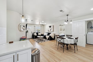 A modern kitchen with white cabinetry, a dining area with a round table, and a living room with a black sofa. - Furnished 4BR Home in Richardson - Extended Stays (Richardson)