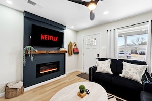 A modern living room with a fireplace, a black sofa with floral cushions, a round coffee table, and a TV mounted above the fireplace displaying 'NETFLIX'. - Furnished 4BR Home in Richardson - Extended Stays (Richardson)