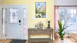 Ein Zimmer mit einer weißen Tür, einem bleiverglasten Dekor, einem Gemälde eines Gebäudes, einem kleinen Tisch mit einer Kerze und einer Blumentopfpflanze.