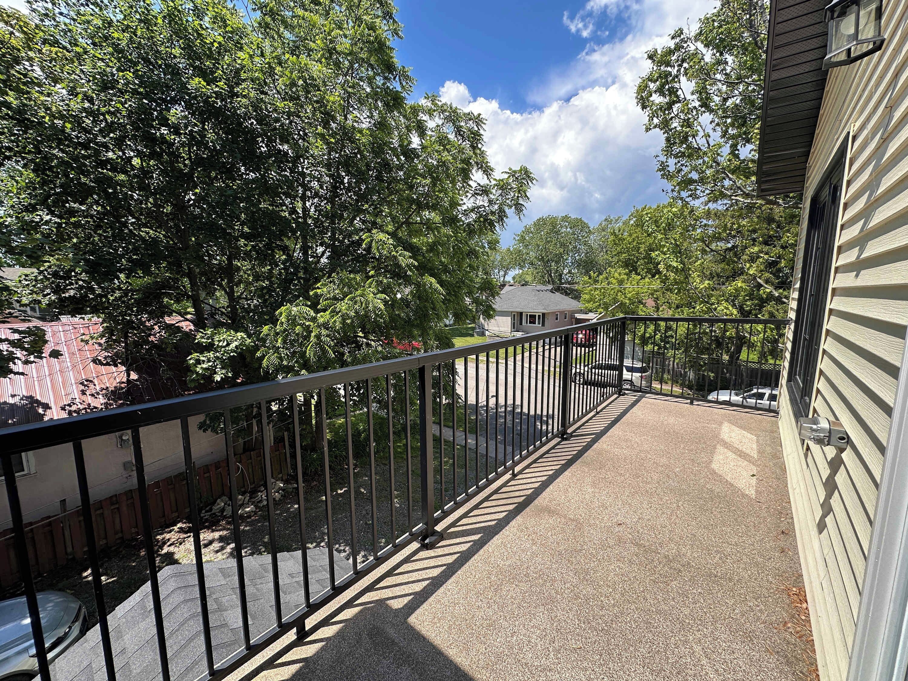 A balcony with a metal railing, a paved walkway, and trees in the background.