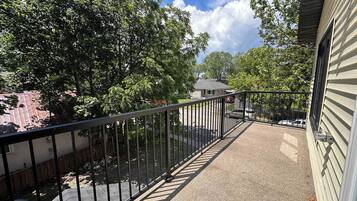 A balcony with a metal railing, a paved walkway, and trees in the background.