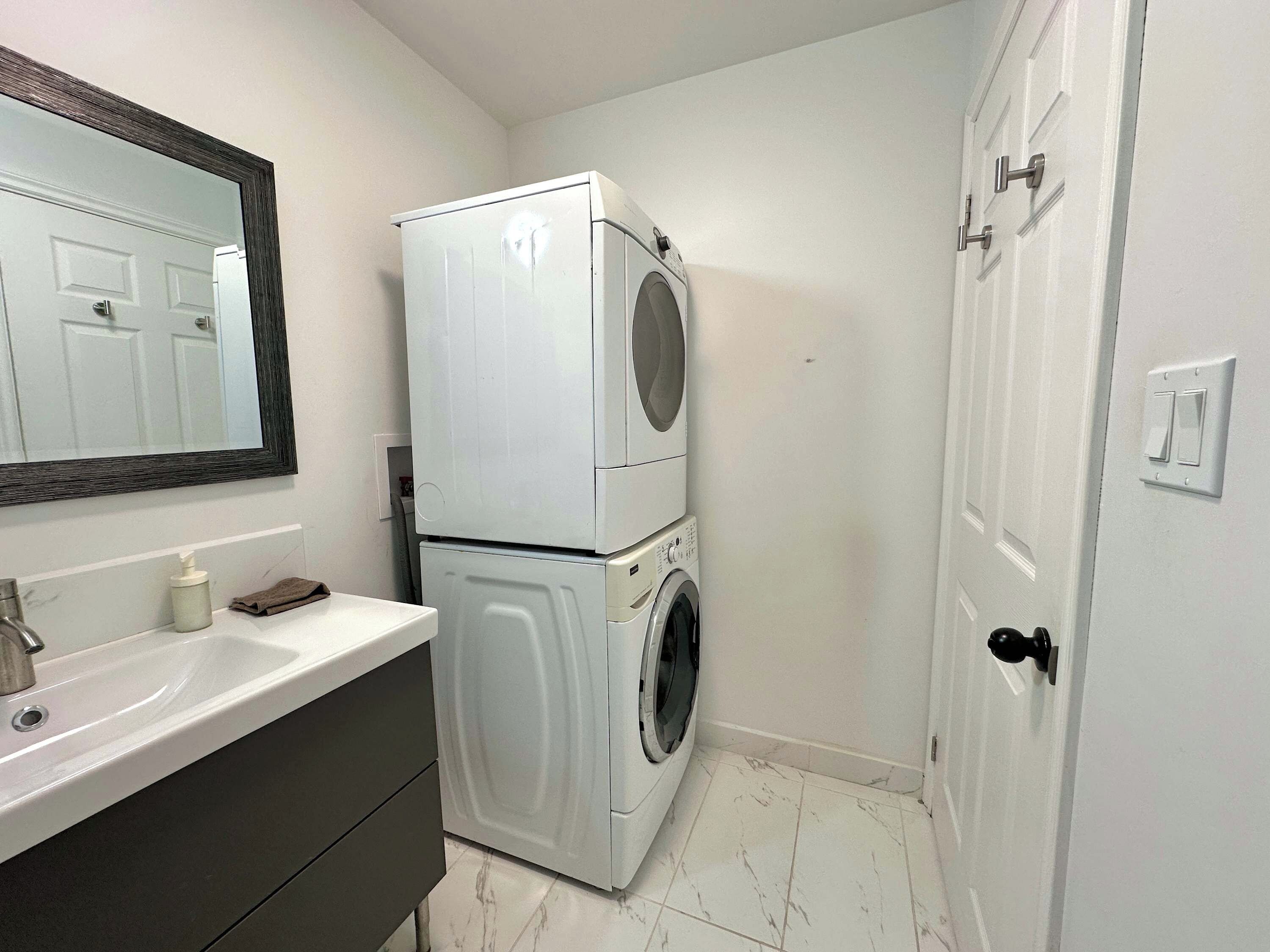 A compact bathroom with a white sink, a mirror, a stacked washer and dryer, and a door with a light switch.
