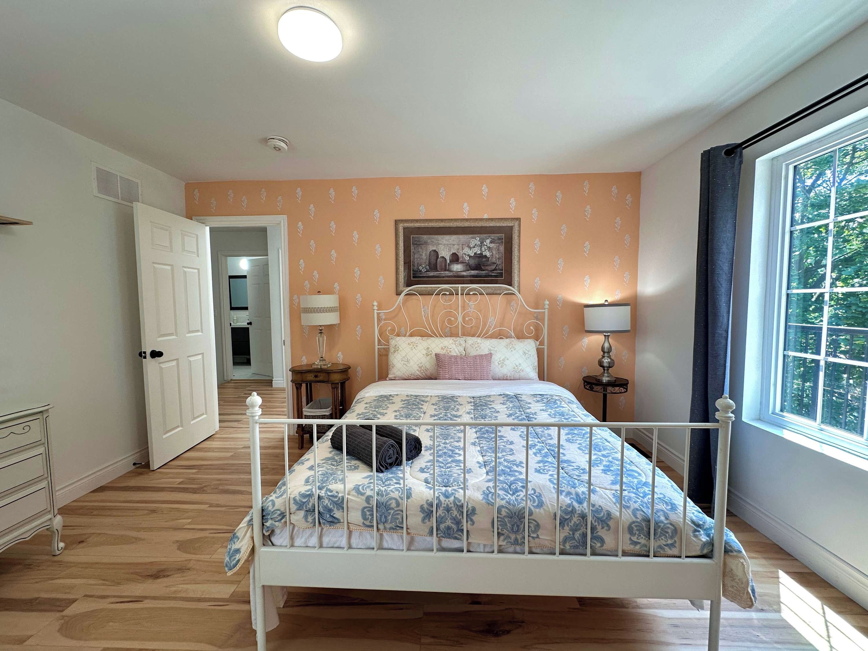 Een slaapkamer met een bed met een wit metalen frame, een bed met een decoratieve spreidek, twee bedlampjes, een raam met gordijnen en een deur naar een andere kamer.