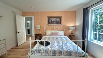Een slaapkamer met een bed met een wit metalen frame, een bed met een decoratieve spreidek, twee bedlampjes, een raam met gordijnen en een deur naar een andere kamer.