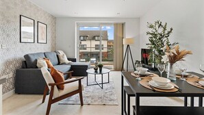 A modern living room with a sofa, a dining table set for two, and a view of buildings through a large window. - Nordic Abode in Central Reading - Balcony (Reading)