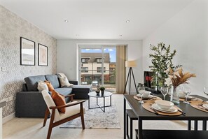 A modern living room with a sofa, a dining table set for two, and a view of buildings through a large window. - Nordic Abode in Central Reading - Balcony (Reading)