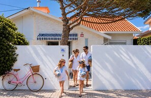 A family walking towards a white building with a tiled roof, a pink bicycle, and a tree.