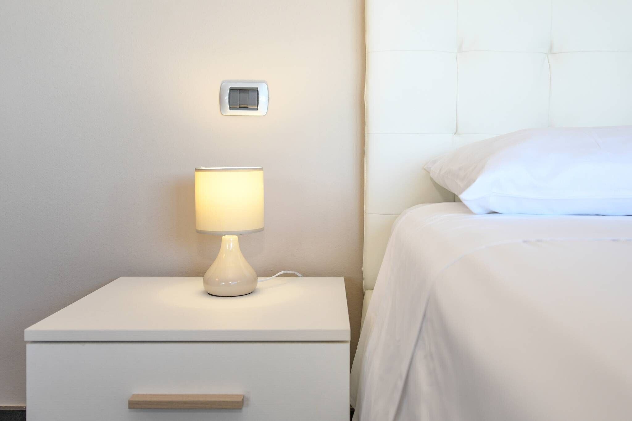 Un lit bien fait, avec du linge de lit blanc, une lampe de chevet, un petit tiroir et un interrupteur fixé au mur.