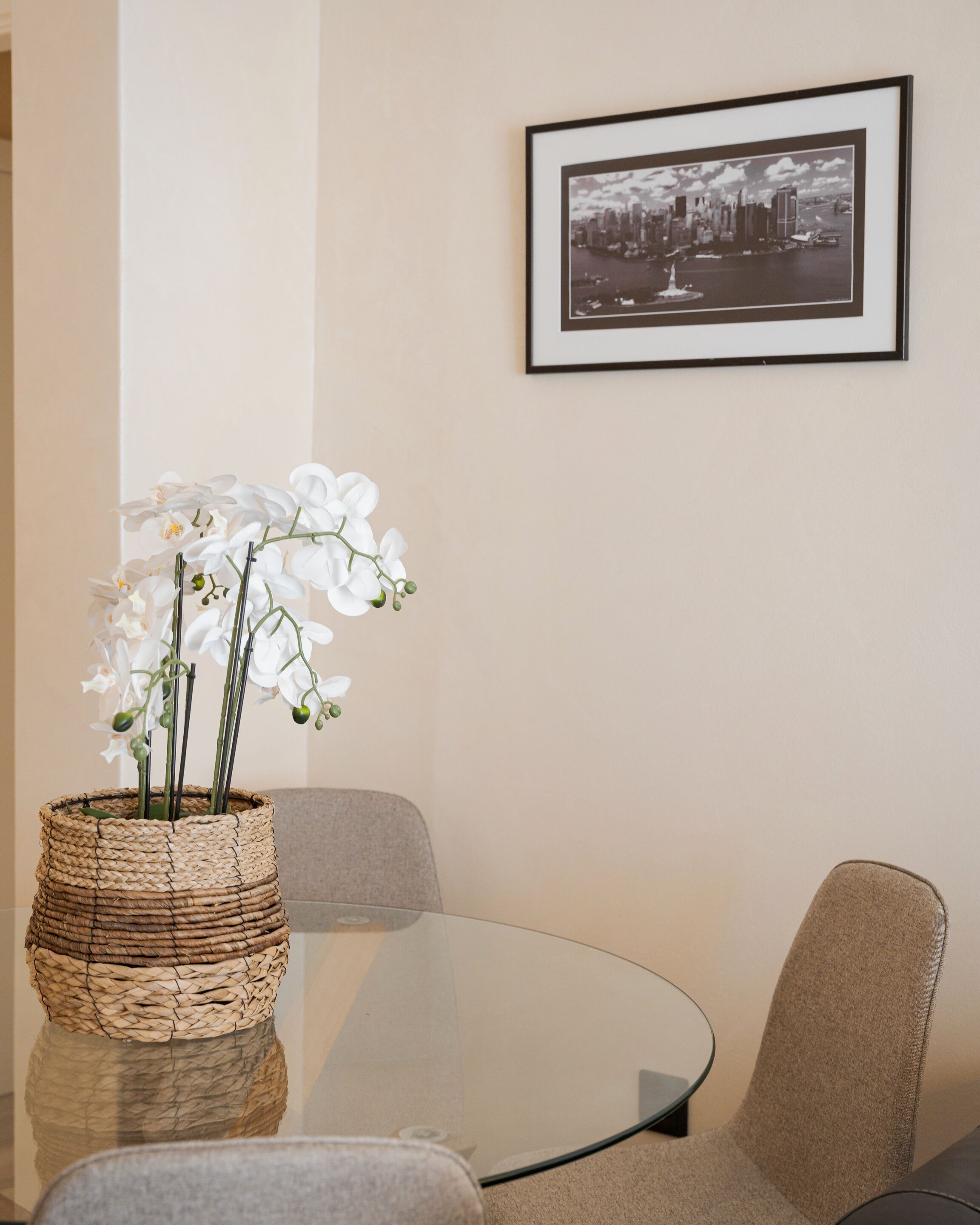 A room with a round glass table, a wicker vase with white flowers, a framed black and white cityscape photo on the wall, and a grey chair.
