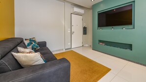 Una sala de estar con un sofá, un televisor montado en una pared color turquesa y una puerta con un equipo de aire acondicionado.