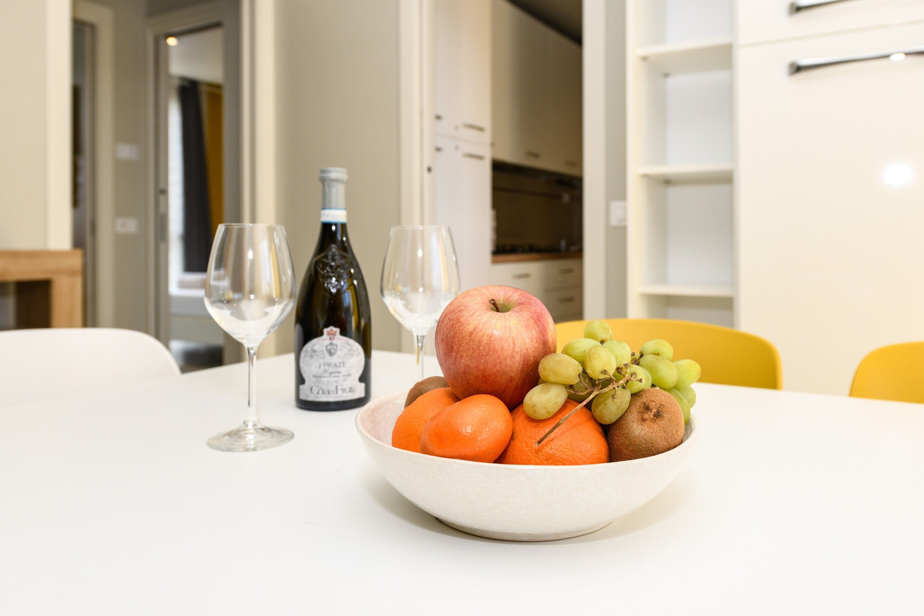 A dining table with a bowl of fruit, two wine glasses, and a bottle of wine.