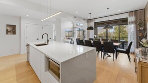 A modern kitchen with a large island, dining area with chairs, and a view of the outdoors. - Mountain Luxe | 4BDR Verbier | Jacuzzi & Sauna (Mont-Tremblant)