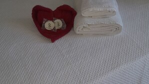A bed with a white textured bedspread, a red heart-shaped pillow with a tag, and a folded white towel.