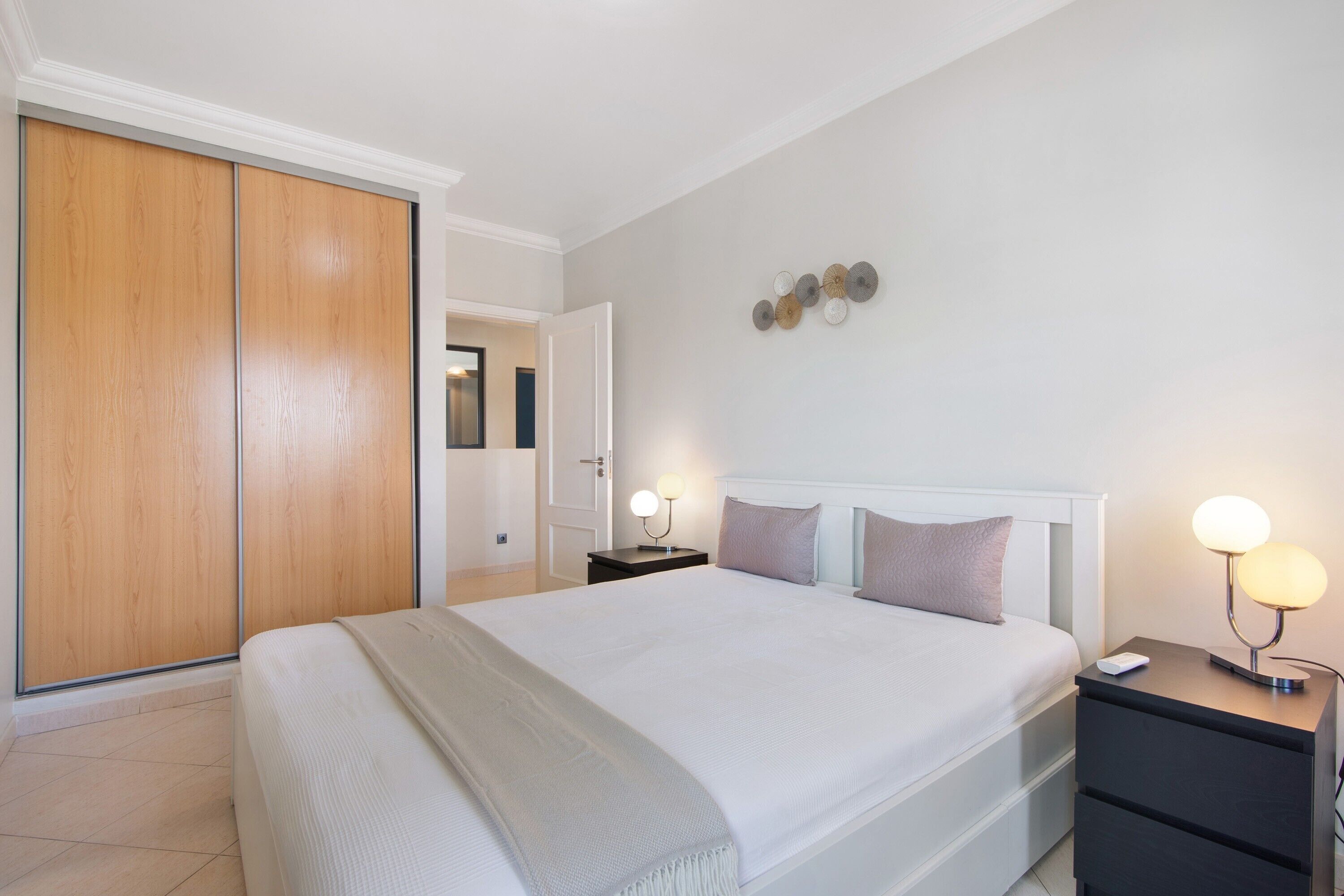 Une chambre avec un grand lit, une armoire, une table de chevet avec une lampe et un luminaire fixé au mur.