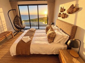 Un dormitorio con una cama grande, una silla colgante, una mesita de noche de madera y vista al océano.