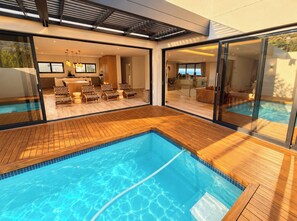 Un área de piscina moderna al aire libre con un deck de madera, un comedor con sillas y vistas al interior a través de puertas de cristal.