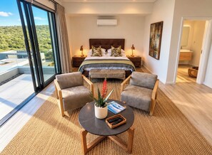 Un dormitorio con una cama grande, dos mesitas de noche, un ventanal con vistas, un cuadro y una zona de estar con una mesa y sillas.