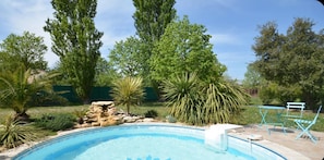 A swimming pool with a rock feature, surrounded by various trees and a green fence. - Charming villa with heated pool Avignon (Saint-Saturnin-lès-Avignon)
