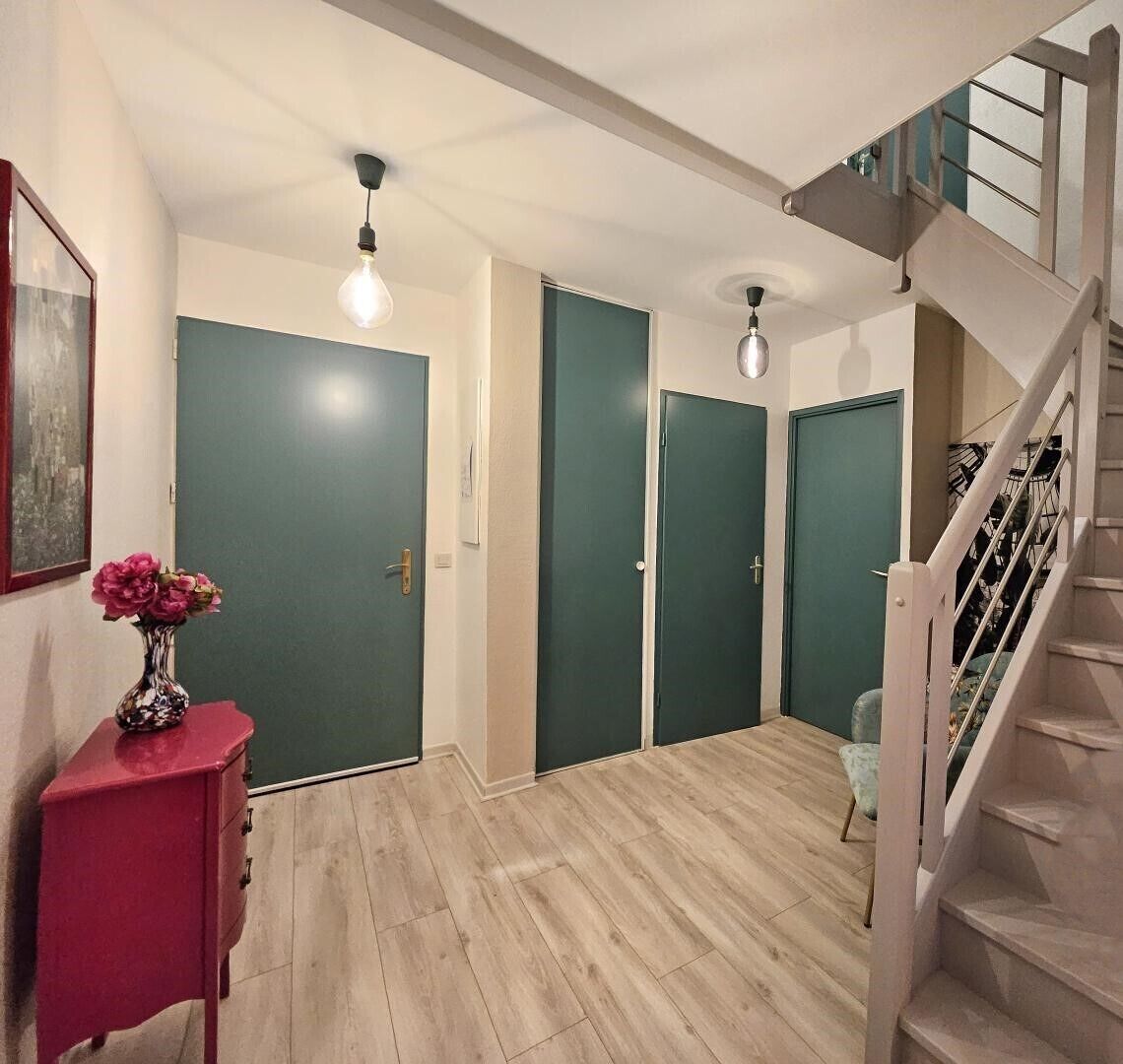 Un couloir doté d’un plancher en bois, de portes vertes, d’un escalier et d’un vase rempli de fleurs posé sur une petite table d’appoint rouge.