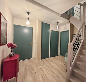 Ein Flur mit Holzboden, grünen Türen, einer Treppe und einer Vase mit Blumen auf einem roten Beistelltisch.