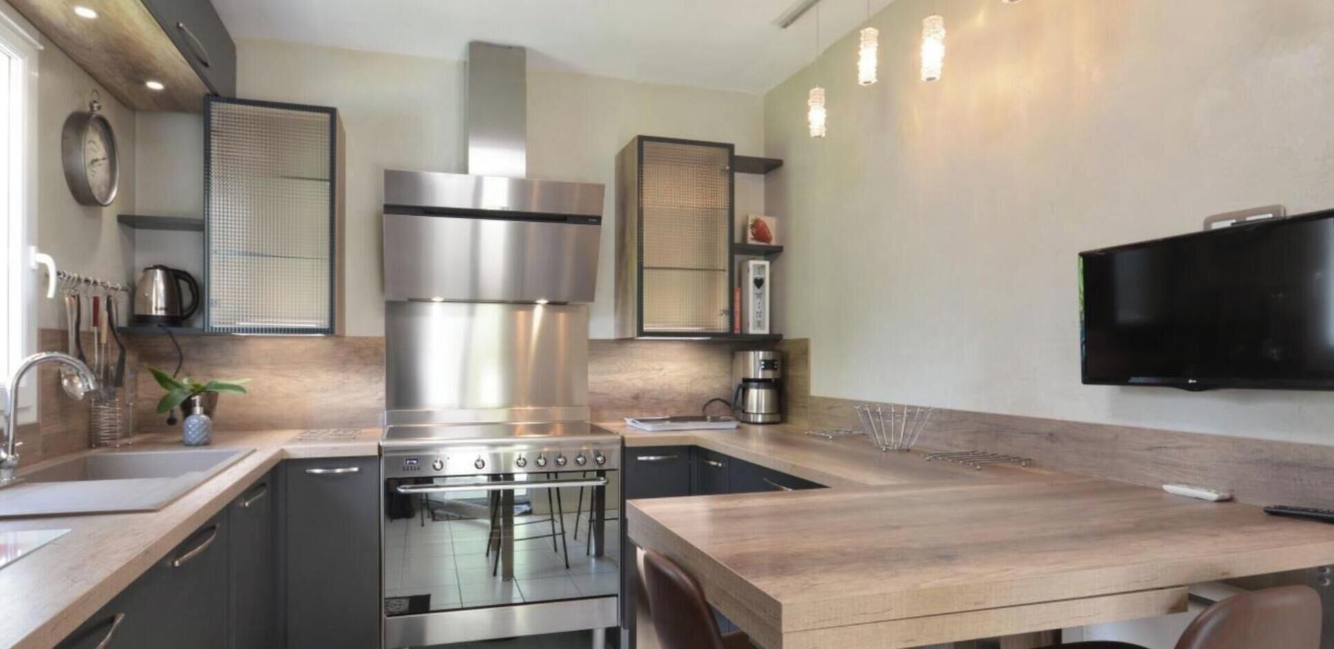 Une cuisine moderne équipée d’une cuisinière, d’un four et d’une machine à café en acier inoxydable. On y trouve une table à manger en bois et un téléviseur fixé au mur.