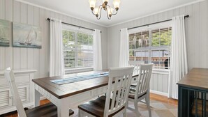 Un comedor con una mesa de madera, sillas blancas, una araña y ventanas con persianas.
