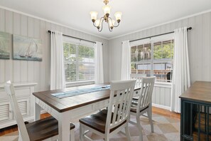 Un comedor con una mesa de madera, sillas blancas, una araña y ventanas con persianas.