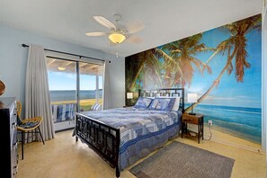 Une chambre à coucher comprenant un lit, un ventilateur de plafond, une table de chevet, une lampe, une chaise, une fenêtre avec des rideaux et une fresque murale représentant une plage.